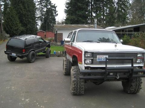 2004 Chevrolet Blazer - Two lifted SUVs are parked in a driveway. The front vehicle appears to be from the 1980s.