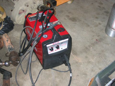 1992 Ford Mustang - A red welding machine sits on a concrete floor, connected to a power source and with its cables and clamps resting on top.