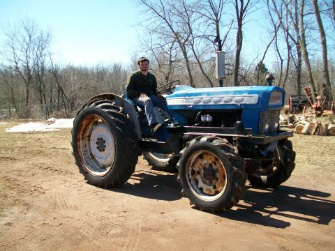 2000 Chevrolet Tahoe - A man sits on a blue tractor in a rural setting with trees and logs visible.