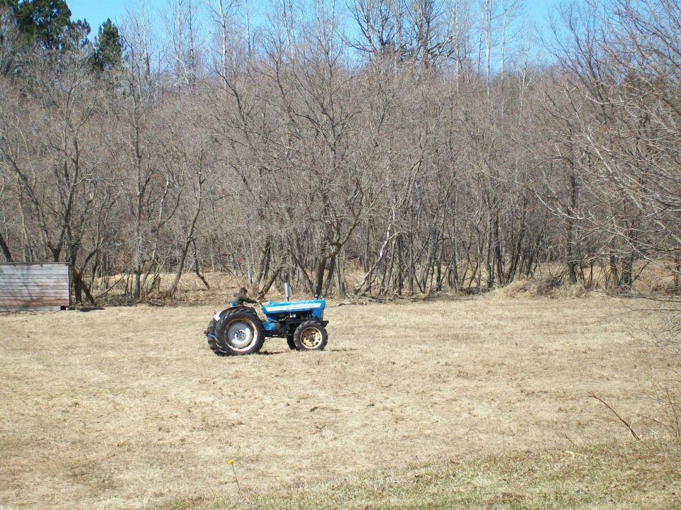 Blue tractor with large rear wheels and smaller front wheels, driven by a person, in a dry, grassy field with bare trees