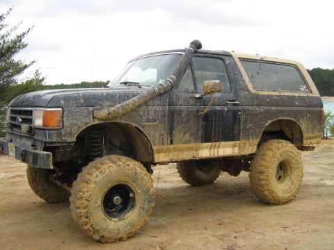 1990 Ford Bronco - A muddy, dark blue, lifted 1990 Ford Bronco is parked on a dirt road with a body of water and trees in the background. The vehicle features…