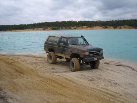 1990 Ford Bronco - A dark colored, lifted Ford Bronco is parked on a sandy beach next to a bright turquoise body of water. The vehicle has large off-road tire…