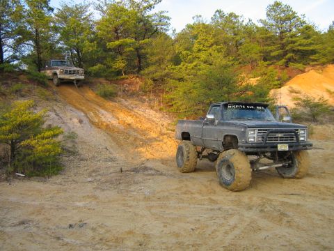 1990 Ford Bronco - Two lifted pickup trucks are parked on a sandy hill with trees in the background. The truck in the foreground is a dark gray Chevrolet pick…