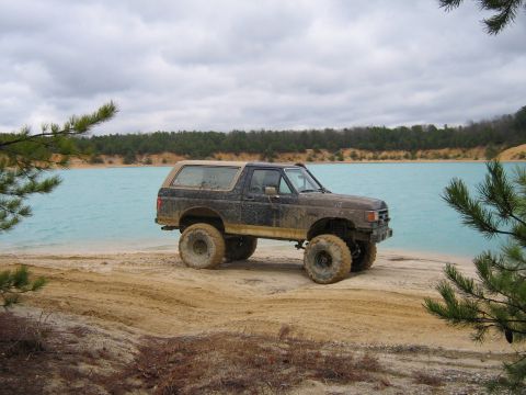 1990 Ford Bronco - A muddy, lifted 1990 Ford Bronco sits on a sandy shore next to a bright turquoise lake.