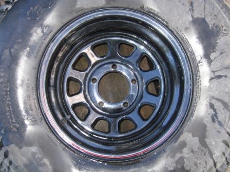1993 Ford Bronco - Close overhead shot of a glossy black slotted steel wagon wheel with a five-lug bolt circle and open center bore, mounted on a whitewall-st…