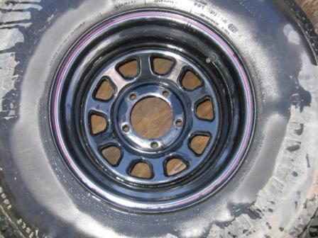 1993 Ford Bronco - Black slotted steel wagon wheel with a five-lug pattern and open center bore, mounted on a red-pinstriped tire and lying face-up on a concr…
