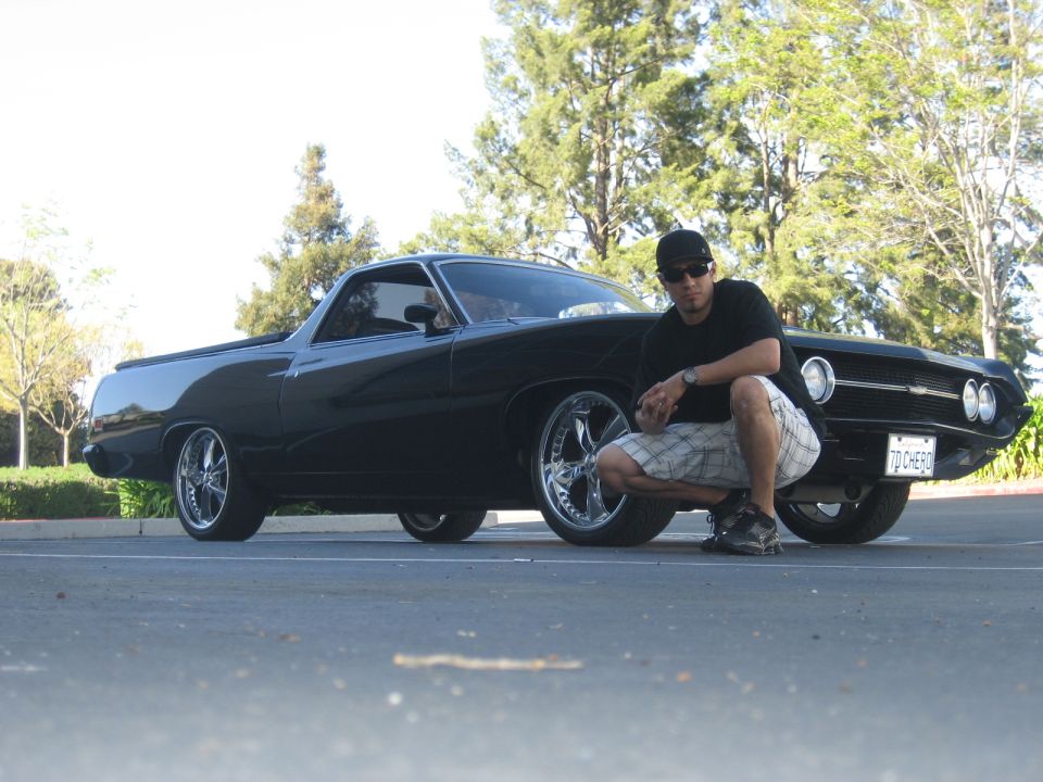 Black Ford Ranchero pickup with chrome wheels, man crouching beside it in parking lot.