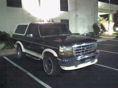 1993 Ford Bronco - A dark colored Ford Bronco is parked in a parking lot at night. The vehicle has a white removable top and white trim along the lower body.