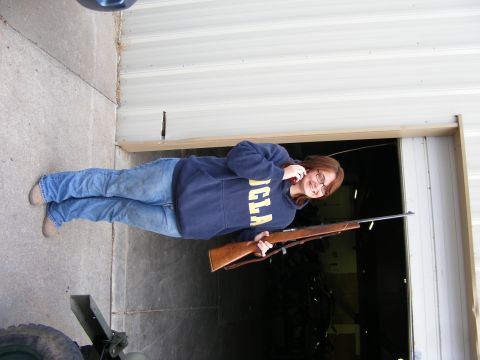 1979 Ford Bronco - A young woman wearing a blue hoodie and jeans holds a rifle and talks on a cell phone while standing in front of an open garage door.