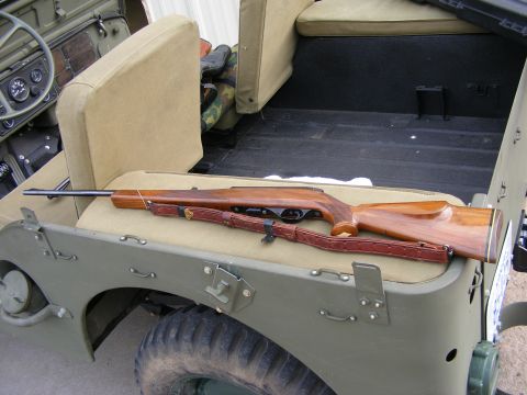 1979 Ford Bronco - A wooden rifle with a leather strap rests on the tan seat of a vintage olive green Jeep. The rifle is positioned horizontally across the se…