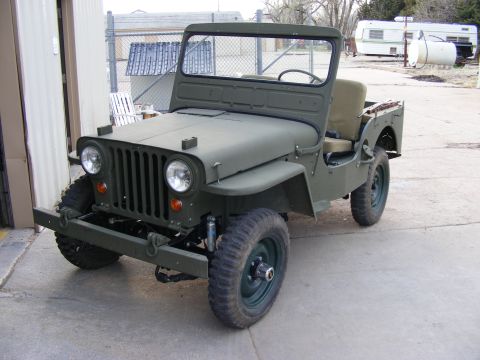 1979 Ford Bronco - A vintage olive green Jeep is parked on a concrete surface in front of a building and a chain-link fence. The Jeep has a spare tire mounted…