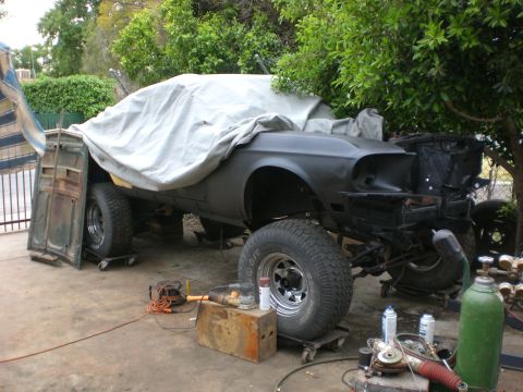 1967 Ford Mustang - A partially covered classic car, likely a Ford Mustang, is undergoing restoration outdoors. The car is elevated on dollies and features lar…