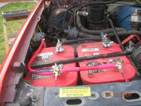 1985 Ford Bronco - The engine bay of a red vehicle shows two red car batteries connected with thick red and pink cables. A yellow sticker with text about refr…