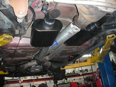 1992 Ford Crown Victoria - The underside of a vehicle is shown, lifted on a yellow hydraulic lift. Visible components include the exhaust system with a muffler, the d…
