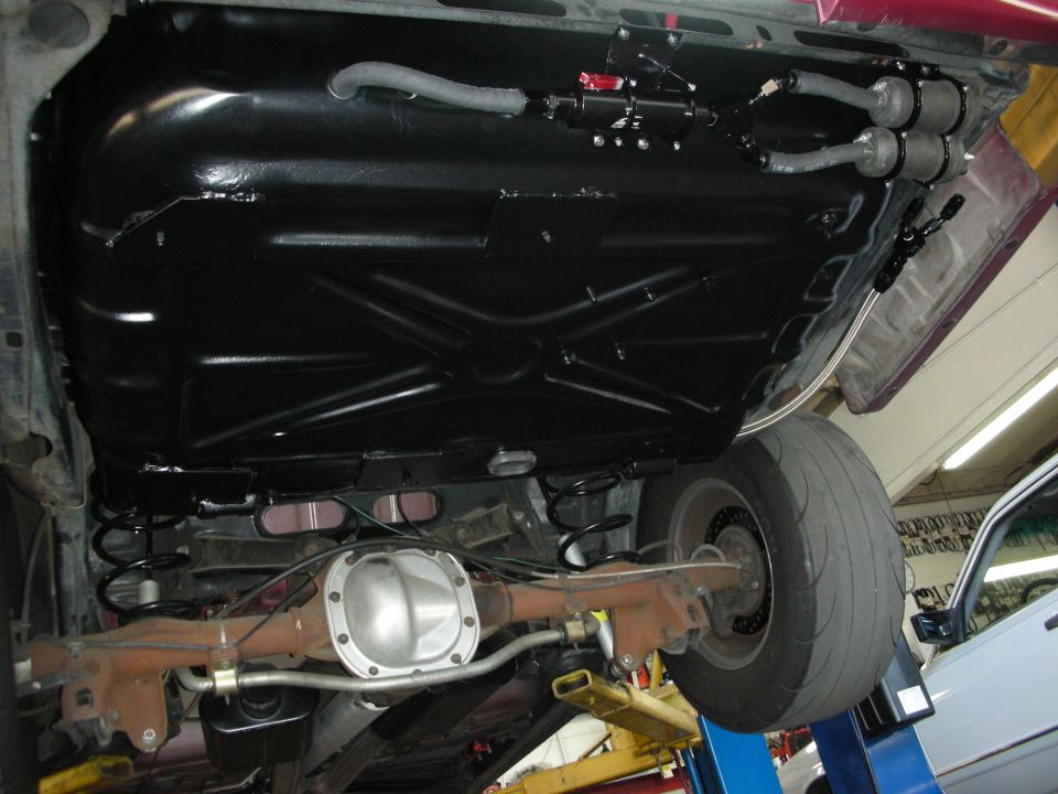 Underside of a car on a lift, showing a black fuel tank, rear axle, and suspension components