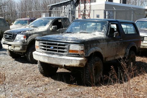 1995 Ford Bronco - Several muddy trucks are parked in a lot with some industrial equipment in the background.