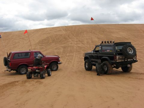 1994 Ford Bronco - Two Ford Broncos and an ATV are parked on sand dunes under a cloudy sky. Both Broncos have flags attached.
