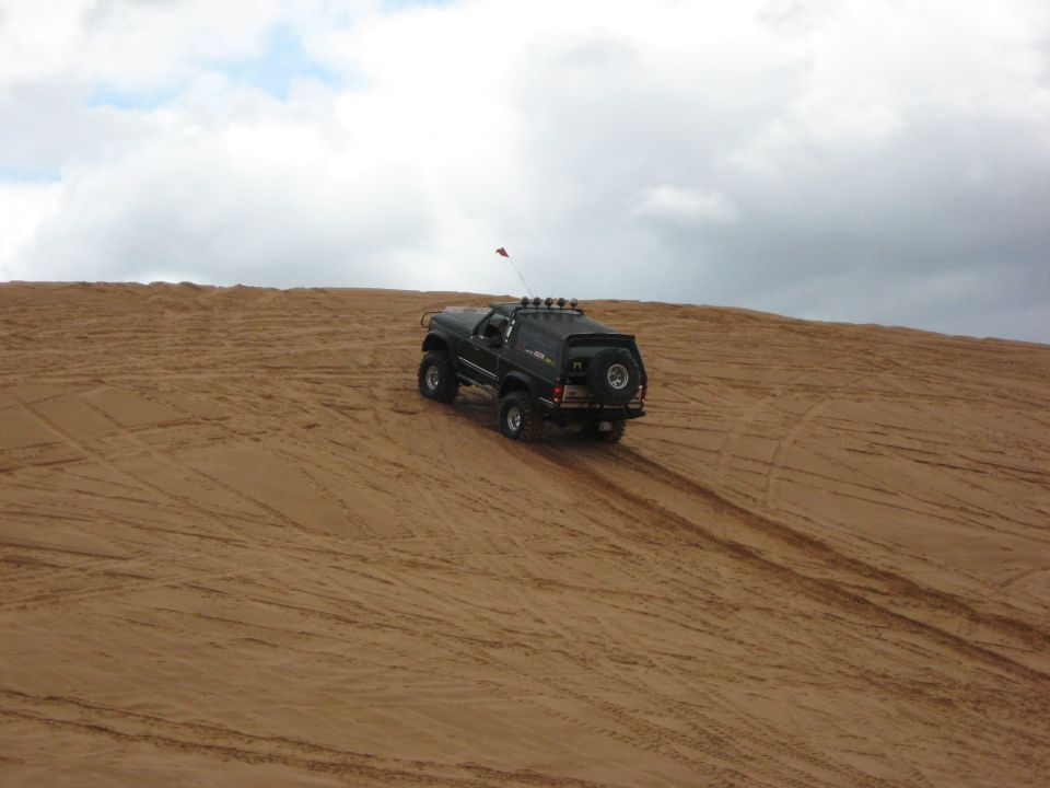 Black SUV with a spare tire on the back driving up a sand dune under a cloudy sky