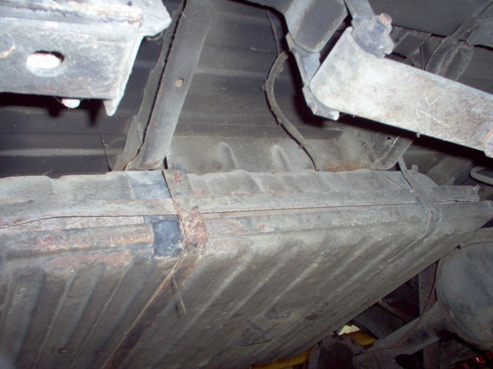 Underside close-up of rusty ribbed floor pan and frame rail with welded seam and body mount
