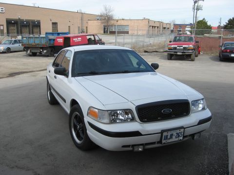 2003 Ford Crown Victoria - A white 2003 Ford Crown Victoria sedan is parked in a lot with other vehicles and buildings in the background.