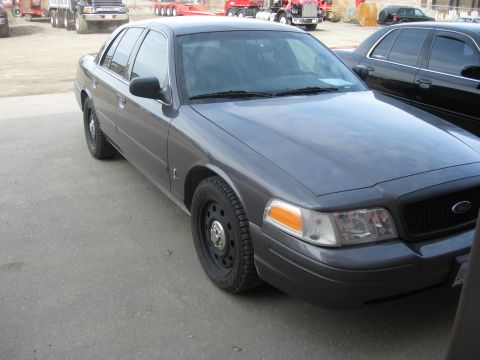 2003 Ford Crown Victoria - A gray 2003 Ford Crown Victoria is parked next to another dark sedan, with trucks in the background.