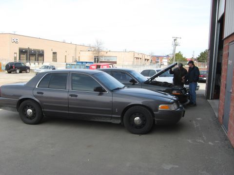 2003 Ford Crown Victoria - A dark gray 2003 Ford Crown Victoria sedan is parked next to another vehicle with its hood open.