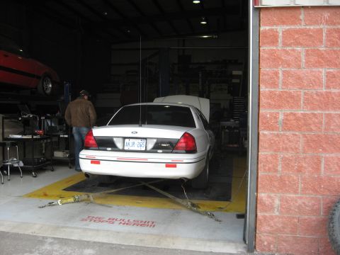 2003 Ford Crown Victoria - A white 2003 Ford Crown Victoria is positioned on a dynamometer in a garage, with another vehicle elevated on a lift in the background.
