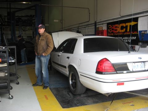 2003 Ford Crown Victoria - A white Crown Victoria sedan with black steel wheels and an Ontario plate sits on a chassis dyno pad inside a shop, hood raised, viewed fro…