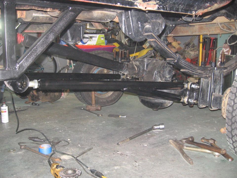 Undercarriage with black suspension links, leaf spring, and axle over garage floor with scattered tools
