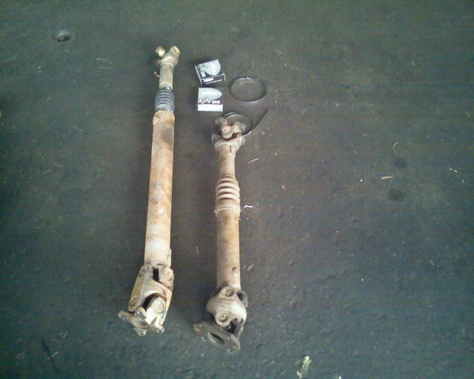 Two rusty driveshafts with U-joints and a spring boot, one longer than the other, on a dark concrete floor