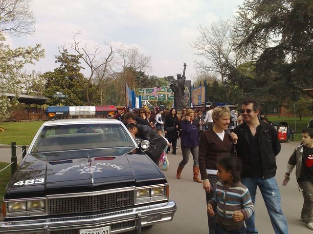 Police car with 'HIGHWAY PATROL' on hood, people in background near a statue
