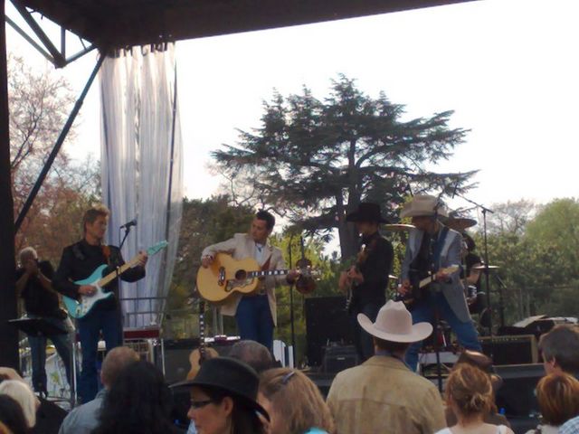 Band playing guitars and singing on a stage outdoors with trees in the background