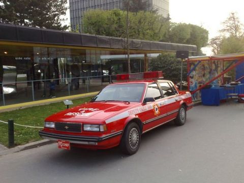 1991 Ford Crown Victoria - This red sedan, marked as a fire department vehicle, is parked near a building with a light bar on its roof.
