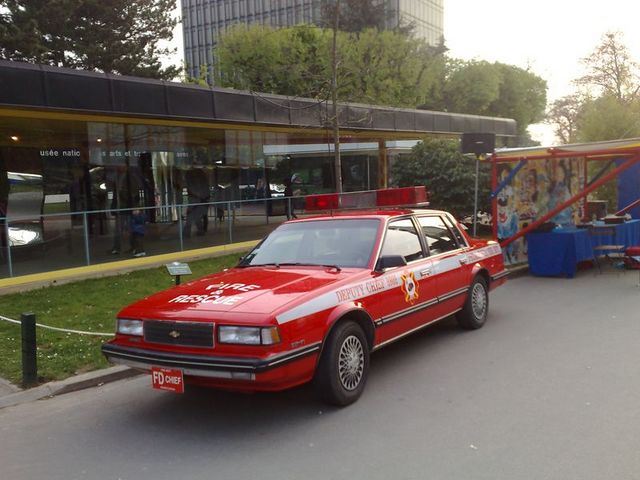 Red fire department car with "FIRE RESCUE" and "DEPUTY CHIEF 100" text, light bar on roof, parked outside a building