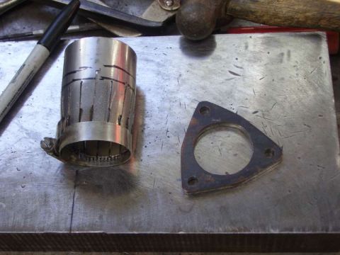 1969 MINI Cooper - A metal adapter for an exhaust pipe sits next to a triangular flange gasket on a metal surface.