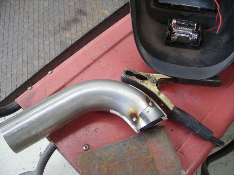 Welding clamp attached to a bent metal pipe, with a welding mask and batteries in the background