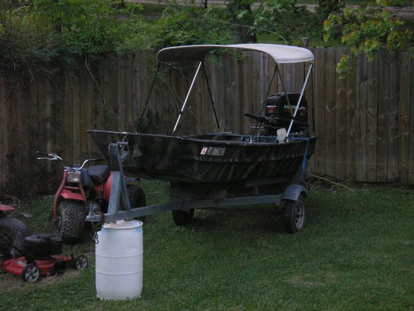 Boat with canopy and Mercury motor on a trailer, parked on grass next to a red ATV and a lawnmower