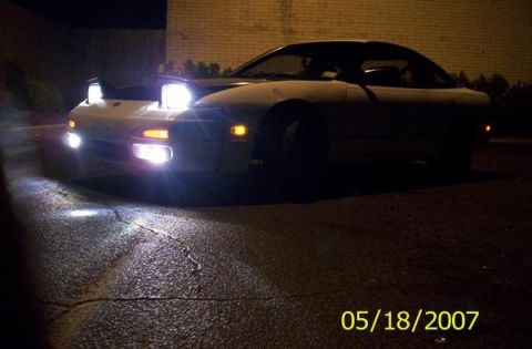1991 Nissan 240SX - A white coupe with its distinctive pop-up headlights illuminated is seen from a three-quarter front view at night.