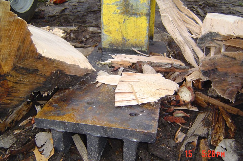 Log splitter steel base plate and yellow wedge amid split wood and bark