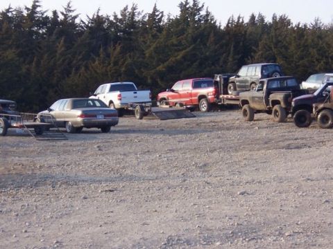 1979 Ford Bronco - A collection of trucks and SUVs are parked on a gravel lot, with a line of trees behind them.
