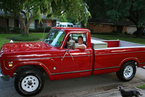1975 International Scout II - A man sits in a red 1975 International pickup truck on a suburban street.