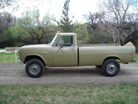 1975 International Scout II - Side profile of a tan International pickup truck, likely from the 1970s, parked on a dirt road with trees in the background.