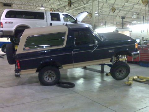 1990 Ford Bronco - A lifted 1990 Ford Bronco is shown on a lift in a garage, with a white pickup truck behind it.