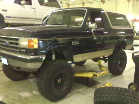 1990 Ford Bronco - A lifted Ford Bronco is being serviced in a garage, with another Bronco visible in the background.