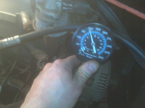1995 Ford Bronco - A hand holds a compression tester gauge with a reading between 100 and 120 psi. The gauge is positioned near engine components, suggesting…