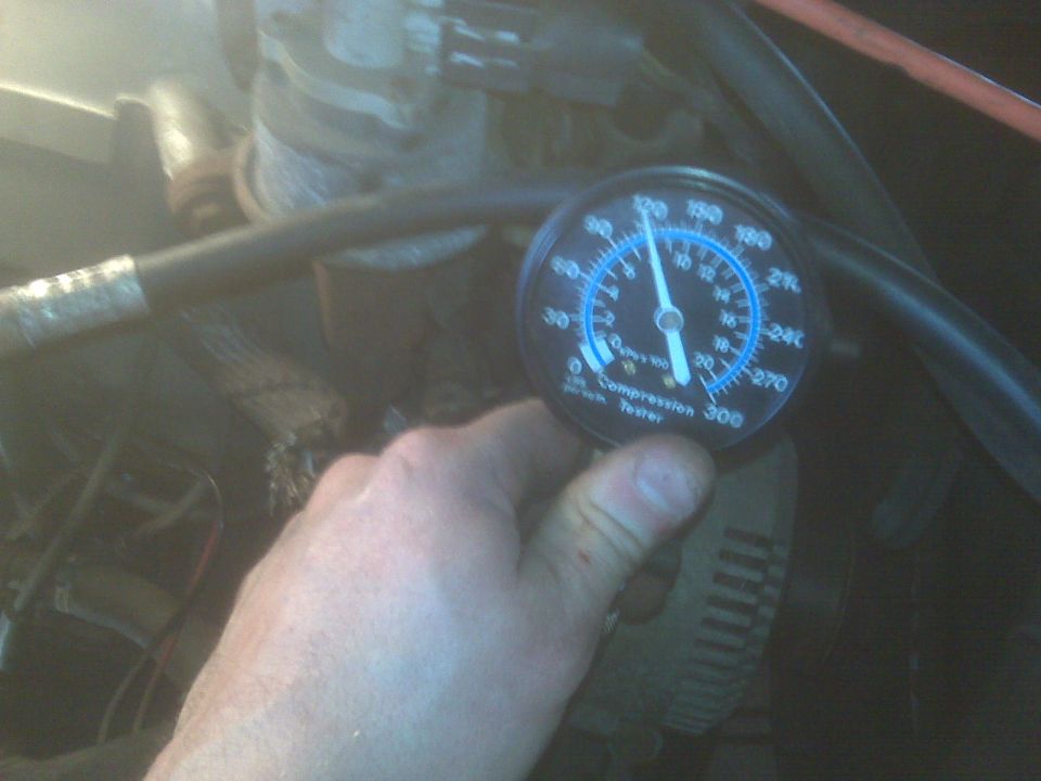 Compression tester gauge showing between 100-120 psi, held near engine parts.