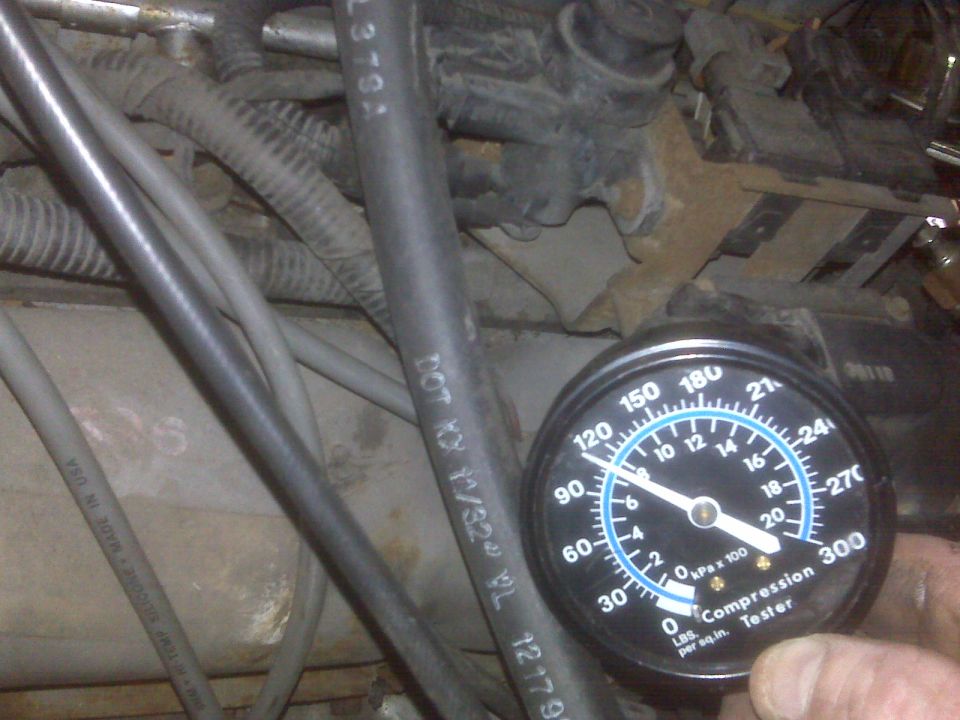 Compression tester gauge showing approximately 110 PSI in front of engine bay components.
