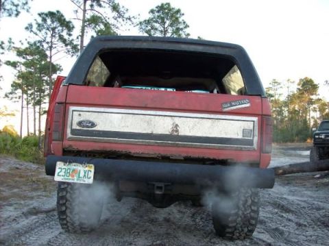 1986 Ford Bronco - The rear driver's side of a red Ford Bronco is shown with smoke coming from the exhaust pipe. A license plate reading "238 LXC" is visible.