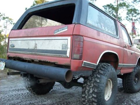1986 Ford Bronco - The rear driver's side quarter view of a red 1980s Ford Bronco is shown. A large, black exhaust pipe extends from beneath the vehicle, and…