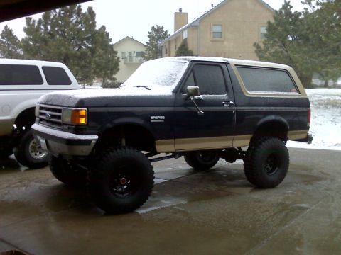 1990 Ford Bronco - A lifted, dark blue and tan Ford Bronco with large tires sits on a wet driveway during a snowfall. A white pickup truck is partially visibl…